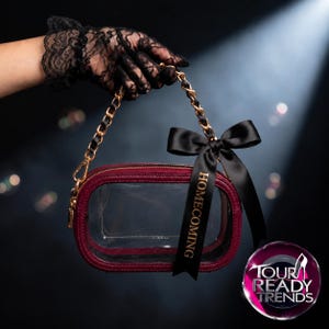 Puede incluir: Un bolso rectangular transparente con un borde texturizado de color rojo oscuro y una correa de cadena dorada. Una cinta de sat&eacute;n negro est&aacute; atada a la correa, con la palabra HOMECOMING impresa en oro. El bolso tiene un cierre de cremallera.