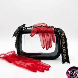 Puede incluir: Un bolso rectangular de pl&aacute;stico transparente con bordes negros, una correa de cadena negra y una cinta negra con la palabra HOMECOMING. Se acompa&ntilde;a de un par de guantes rojos transparentes, uno colocado encima y otro delante del bolso.