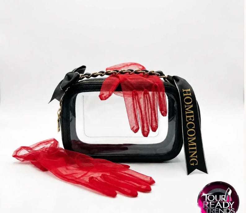 Puede incluir: Un bolso rectangular de pl&aacute;stico transparente con bordes negros, una correa de cadena negra y una cinta negra con la palabra HOMECOMING. Se acompa&ntilde;a de un par de guantes rojos transparentes, uno colocado encima y otro delante del bolso.