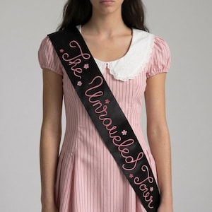 May include: A light pink dress with thin vertical dark stripes, short puff sleeves, and a white scalloped collar. A black sash is worn diagonally across the chest, featuring the embroidered pink text "The Unraveled Sour" and small pink flower accents.