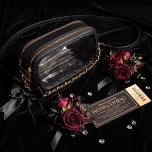 Puede incluir: Un bolso bandolera rectangular negro con panel frontal transparente, borde de cadena dorada y lazo de sat&eacute;n negro. Dos ramilletes de rosas rojo oscuro con cintas negras y una entrada de Homecoming con texto dorado se disponen junto al bolso.