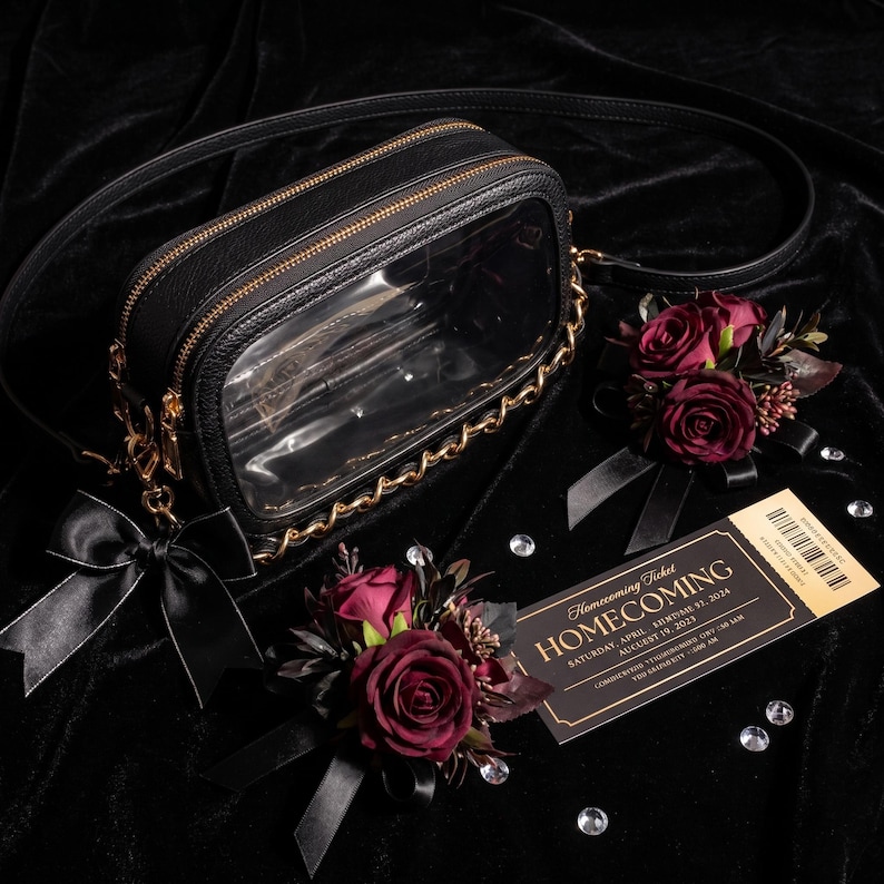 Puede incluir: Un bolso bandolera rectangular negro con panel frontal transparente, borde de cadena dorada y lazo de sat&eacute;n negro. Dos ramilletes de rosas rojo oscuro con cintas negras y una entrada de Homecoming con texto dorado se disponen junto al bolso.
