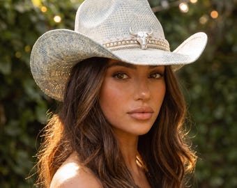 May include: Cowboy hat with a light blue and white pattern. The hat features a silver-colored bull skull emblem and a decorative band. The brim is curved, and the hat has a classic western style. The hat is made of a woven material.