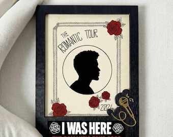 May include: A black framed art piece featuring a silhouette of a person's profile inside a circle, with the text "THE ROMANTIC TOUR" above. Red roses and the text "2024" are also present. The bottom of the frame reads "I WAS HERE THE ROMANTIC TOUR 2026".