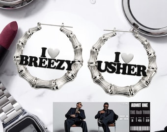 May include: A pair of silver-toned bamboo-style hoop earrings. One earring features the black text "I [heart symbol] BREEZY" and the other features "I [heart symbol] USHER" in black lettering. The earrings are displayed alongside a digital watch.