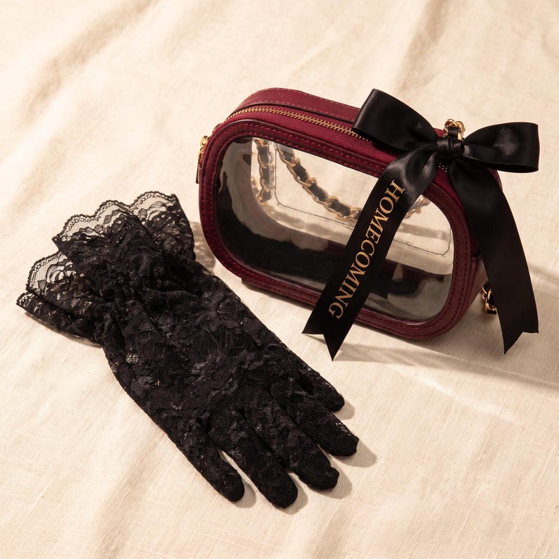 Puede incluir: Un bolso ovalado transparente con borde rojo oscuro, correa de cadena dorada y un lazo de sat&eacute;n negro con el texto HOMECOMING. Junto al bolso hay un par de guantes de encaje negro con bordes festoneados y patrones florales intrincados.