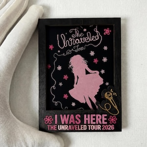 Puede incluir: Un letrero rectangular de madera negra con la silueta rosa de una persona con vestido, rodeada de pequeñas flores rosas. El texto dice "The Unraveled Tour" y "I WAS HERE THE UNRAVELED TOUR 2026" con un icono de micrófono dorado.