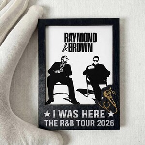 Puede incluir: Un marco rectangular de madera negra que contiene una impresión blanca con el texto RAYMOND & BROWN sobre dos figuras sentadas. Debajo, el texto I WAS HERE THE R&B TOUR 2026 está flanqueado por dos estrellas y un icono de micrófono dorado.