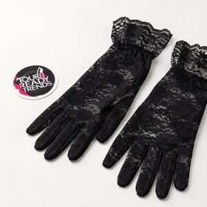 Puede incluir: Un par de guantes de encaje negro con un patr&oacute;n floral y un pu&ntilde;o con volantes en la mu&ntilde;eca. Junto a los guantes hay una pegatina circular con el texto TOUR READY TRENDS y un gr&aacute;fico de un zapato de tac&oacute;n en blanco, rosa y negro.