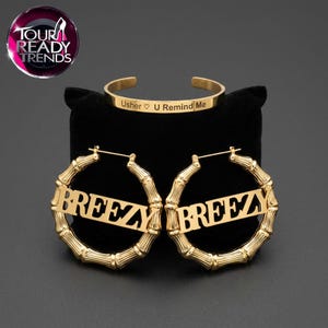 May include: A gold-toned jewelry set consisting of a cuff bracelet and two hoop earrings. The bracelet is engraved with the text Usher ♡ U Remind Me. The hoop earrings feature a bamboo-style texture and the word BREEZY centered in the middle.
