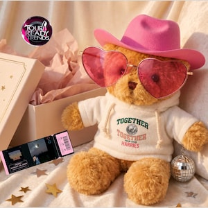 Puede incluir: Un oso de peluche color canela con un sombrero de vaquero rosa brillante y gafas de sol rojas en forma de corazón. El oso lleva una sudadera blanca con el texto TOGETHER TOGETHER TOUR 2026 HARRIES impreso. Una pequeña bola de discoteca plateada cuelga del oso.