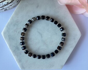 Puede incluir: Una pulsera de cuentas negras con cuentas cuadradas y espaciadores plateados. La pulsera se exhibe sobre una superficie hexagonal de mármol blanco. Las cuentas tienen una apariencia facetada, reflejando la luz. Es una pieza de joyería.