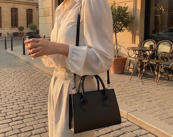 Peut inclure: Une chemise blanche à manches longues boutonnée avec un col et des poignets, associée à un pantalon beige taille haute à plis. Un sac à main rectangulaire noir structuré avec deux anses et une bandoulière est porté à l'épaule.