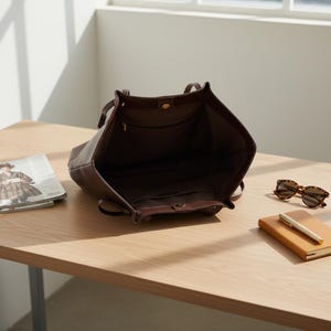 Puede incluir: Bolso tote de cuero marr&oacute;n abierto con asas, que muestra los bolsillos interiores y un cierre a presi&oacute;n. El bolso est&aacute; sobre una mesa de madera junto a una revista, gafas de sol y un cuaderno con un bol&iacute;grafo.
