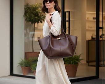 Peut inclure: Une femme portant des lunettes de soleil et une robe crème porte un grand sac fourre-tout en cuir marron foncé avec un fermoir doré. Le sac a deux poignées et un design structuré, adapté à un usage quotidien.