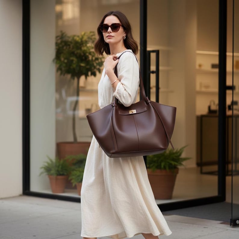 Puede incluir: Una mujer con gafas de sol y un vestido color crema lleva un bolso tote de cuero marr&oacute;n oscuro con un cierre dorado. El bolso tiene dos asas y un dise&ntilde;o estructurado, adecuado para el uso diario.