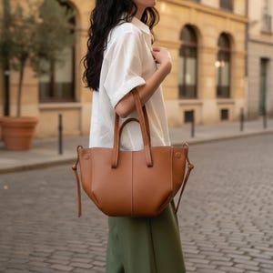 May include: A tan leather tote bag with a structured, curved silhouette, two top handles, and side drawstring details with knotted ends. The bag features a smooth, solid-colored exterior with visible vertical stitching down the center front panel.