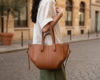 Peut inclure: Un sac cabas en cuir marron clair avec une silhouette incurvée et structurée, deux anses supérieures et des détails de cordon de serrage sur les côtés avec des extrémités nouées. Le sac présente un extérieur lisse avec une couture verticale.