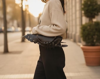 Peut inclure: Un sac à bandoulière en cuir noir en forme de homard, doté de sections de queue segmentées, de petits appendices en forme de pinces et de clous métalliques. Le sac est porté avec un pull beige à manches longues et un pantalon noir en extérieur.