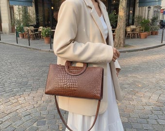 Peut inclure: Un sac à main rectangulaire marron avec une texture gaufrée façon crocodile, doté d'une poignée supérieure circulaire et d'une fine bandoulière. Le sac est porté avec un blazer beige et une robe blanche, affichant un design structuré.