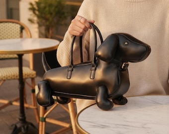 Peut inclure: Un sac à main noir en forme de chien teckel. Le sac présente une finition lisse et mate, un corps structuré avec quatre petites pattes, une longue queue et une tête aux oreilles tombantes. Deux fines anses noires sont fixées sur le dos.