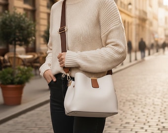 Peut inclure: Un sac seau de couleur crème avec une patte décorative en similicuir marron et une large bandoulière marron réglable dotée de finitions dorées, porté sur un pull en maille côtelée crème et un pantalon sombre.