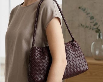 Peut inclure: Un sac à bandoulière bordeaux avec un motif tissé. Le sac a une bandoulière tressée et est porté sur l'épaule. La couleur du sac complète les vêtements aux tons neutres.