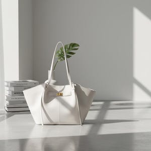 Puede incluir: Un bolso tote de cuero blanco con asas largas y un cierre dorado. La bolsa est&aacute; vac&iacute;a y se encuentra sobre una superficie clara. Un jarr&oacute;n blanco con una hoja verde y libros est&aacute;n en el fondo.