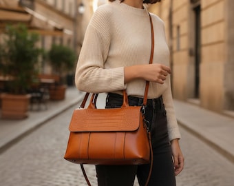 Peut inclure: Un sac à main en cuir marron clair avec un rabat à motif crocodile, deux poignées supérieures et une bandoulière amovible. Le sac présente une base rectangulaire structurée, des coutures verticales sur le devant et des détails en métal foncé.