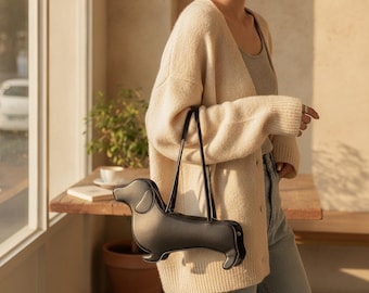 Peut inclure: Un sac à main noir en forme de chien teckel avec une finition lisse, doté de deux fines bandoulières noires fixées sur le dessus. Le sac présente une silhouette canine distincte avec un long corps, des pattes courtes et une tête arrondie.
