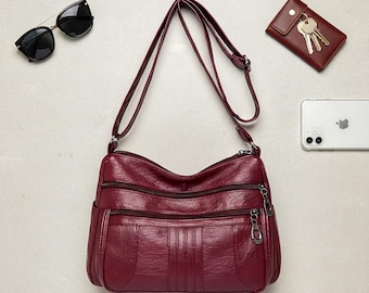 Peut inclure: Un sac bandoulière bordeaux avec plusieurs compartiments zippés et une longue bandoulière. Le sac est fait d'un matériau texturé et possède des accessoires argentés. Sont également présentés des lunettes de soleil noires, un portefeuille bordeaux avec des clés et un smartphone blanc.