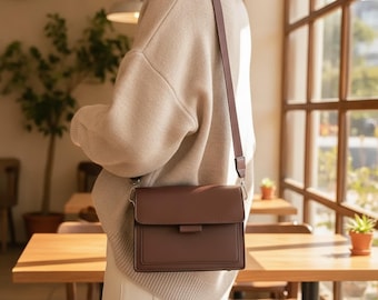 Peut inclure: Un sac bandoulière rectangulaire marron uni avec une fermeture à rabat et une bandoulière réglable assortie, porté sur un pull en maille beige. Le sac présente des lignes épurées, une finition lisse et des attaches en métal argenté.