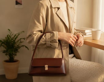 Peut inclure: Un sac à bandoulière rectangulaire marron foncé à la finition lisse, doté d'une poignée supérieure unique et d'un fermoir métallique doré sur le rabat avant. Le sac présente une silhouette structurée et est porté avec une tenue aux tons neutres.