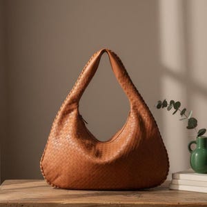 May include: A tan leather hobo bag with a woven design. The bag has a single shoulder strap and a zipper closure. The bag is sitting on a wooden surface.