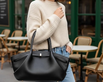 Peut inclure: Un sac fourre-tout en cuir noir avec un design structuré. Le sac a deux poignées et un fermoir doré. Le sac est porté par une personne vêtue d'un pull crème et d'un jean bleu.