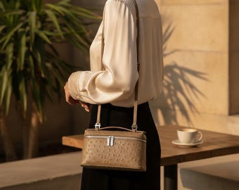 Peut inclure: Un sac à main rectangulaire taupe avec un motif texturé façon autruche, doté d'une poignée supérieure, d'une fine bandoulière réglable et d'une fermeture éclair argentée sur le dessus. Le sac présente une forme structurée et rigide.