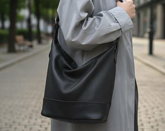 Peut inclure: Un sac à bandoulière en cuir noir avec une base rectangulaire et une longue sangle. Le sac est porté à l'épaule. L'image est prise à l'extérieur. Le sac semble être fait d'un cuir lisse et mat.