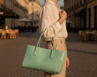 Peut inclure: Un sac cabas vert menthe avec un motif texturé gaufré façon crocodile, doté de deux anses structurées et d'une silhouette rectangulaire, porté sur le côté d'une chemise blanche à manches longues et d'un pantalon beige.