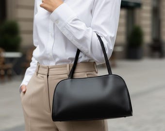 Peut inclure: Un sac à main noir de forme rectangulaire avec le dessus incurvé et deux fines poignées. La personne porte une chemise blanche et un pantalon beige. L'arrière-plan est flou, suggérant un cadre extérieur.