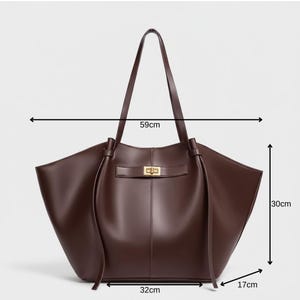 Puede incluir: Un bolso tote de cuero marr&oacute;n con forma rectangular. El bolso tiene un cierre dorado y dos asas largas. El bolso mide 59 cm de ancho, 30 cm de alto y 17 cm de profundidad.