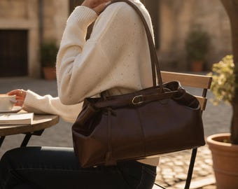 Peut inclure: Un sac à épaule en cuir marron foncé doté d'une sangle horizontale sur le devant avec une boucle en métal doré. Le sac présente une base structurée, une finition lisse et deux longues bandoulières, dans une teinte marron chocolat profond.