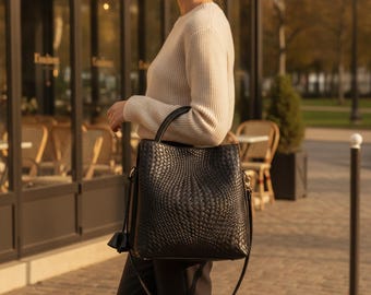 Peut inclure: Un sac seau noir avec une texture en cuir tressé, une base structurée et une longue bandoulière. Le sac présente une ouverture supérieure lisse et un petit pompon décoratif suspendu sur le côté, offrant un design épuré et fonctionnel.