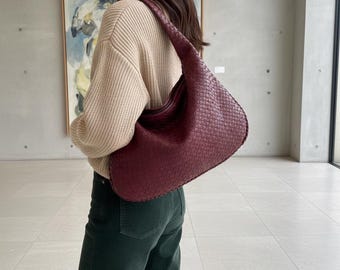 Peut inclure: Un sac à bandoulière bordeaux tissé avec un design incurvé. Le sac est fait d'un matériau texturé et possède une longue sangle. Le sac est porté sur l'épaule. L'arrière-plan comprend un pull beige et un pantalon vert.