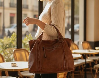 Peut inclure: Un grand sac cabas en cuir marron doté d'accents tressés sur les côtés, d'une poche frontale zippée avec une longue tirette et de deux anses robustes. Le sac présente une base rectangulaire structurée et des coutures visibles sur les côtés.