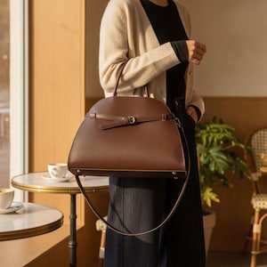 May include: A structured dark brown handbag with a semi-circular top, featuring a thin horizontal strap across the front with a small metal buckle, a top handle, and a long, thin shoulder strap that hangs down in a large loop.