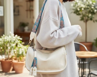 Peut inclure: Sac à bandoulière crème avec une bandoulière décorative à motifs. Le sac a une forme incurvée, une fermeture éclair et un détail de pompon. La bandoulière présente un motif bleu, orange et blanc. Le sac est porté à l'épaule.