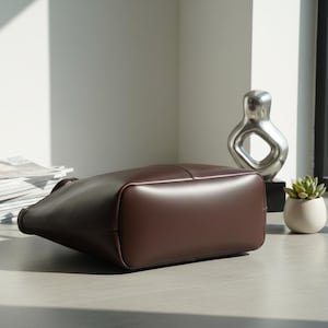 Puede incluir: Un bolso tote de cuero marr&oacute;n oscuro con base rectangular. La bolsa est&aacute; sobre una superficie gris claro, con una escultura plateada y una peque&ntilde;a suculenta en maceta en el fondo.