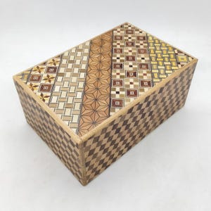 May include: A rectangular wooden puzzle box with intricate geometric patterns. The top features various mosaic designs, including stars, squares, and diamonds, in shades of brown, beige, and yellow. The sides have a diamond pattern.