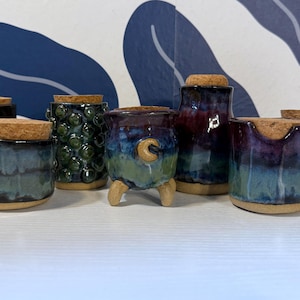 May include: A selection of handmade ceramic jars with cork stoppers. The jars are diverse in shape, size, and colour, featuring blue, green, and purple tones. Some have textured surfaces, while others are smooth. The jars are arranged on a white surface.