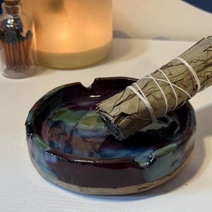 May include: A handmade ceramic ashtray with a dark purple, blue, and green glaze. A bundle of dried sage is resting in the ashtray. A small glass bottle with matches and a lit candle are in the background.
